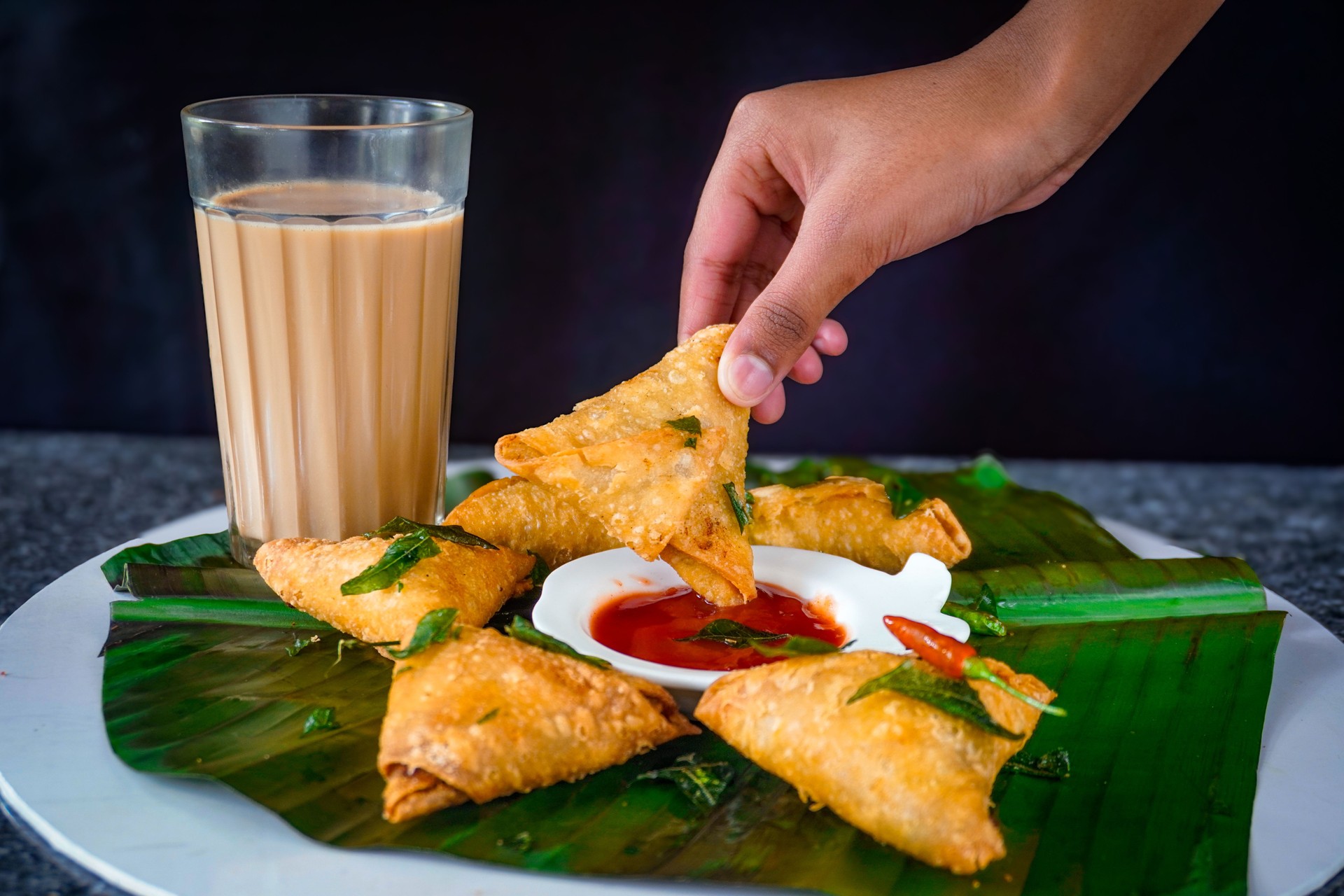 Image of hand dipping samosa into tomato sauce and ketchup with chai tea, banana leaf in sri lankan fast food style